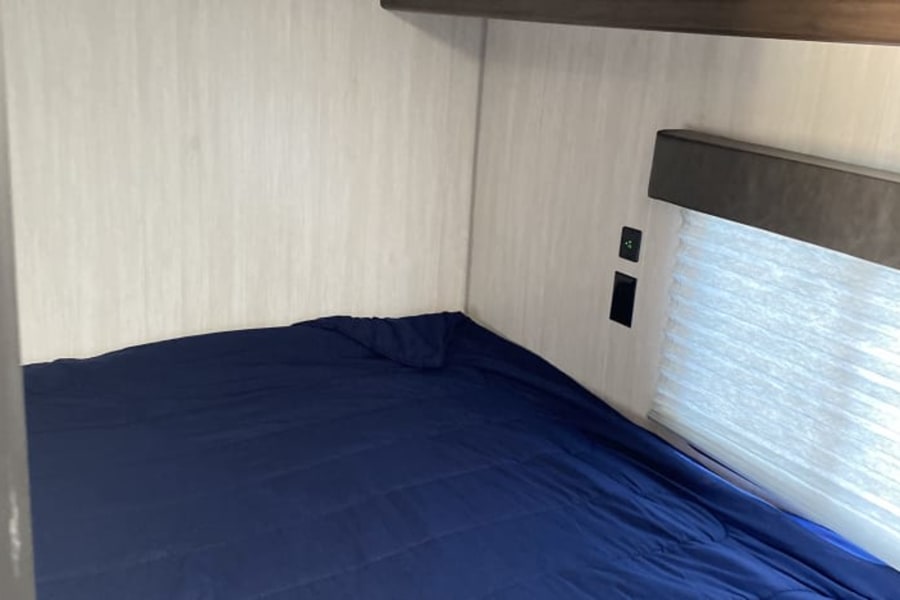 Cozy RV rental bunk bed with a blue comforter, light walls, power outlets, and window blinds. A comfortable sleeping space.