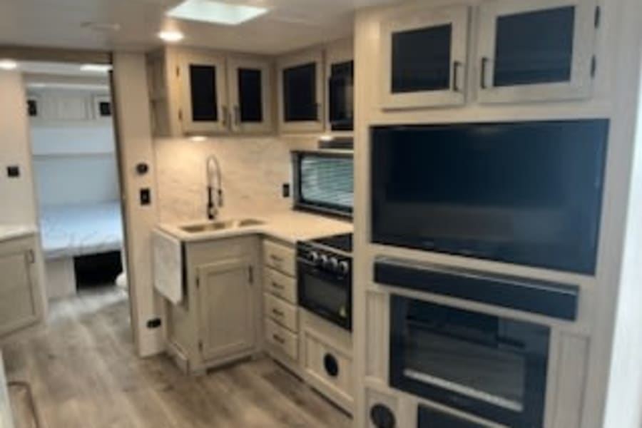 Spacious rv rental interior showing a full kitchen, TV entertainment center with fireplace, and a glimpse of the bedroom. Modern design.