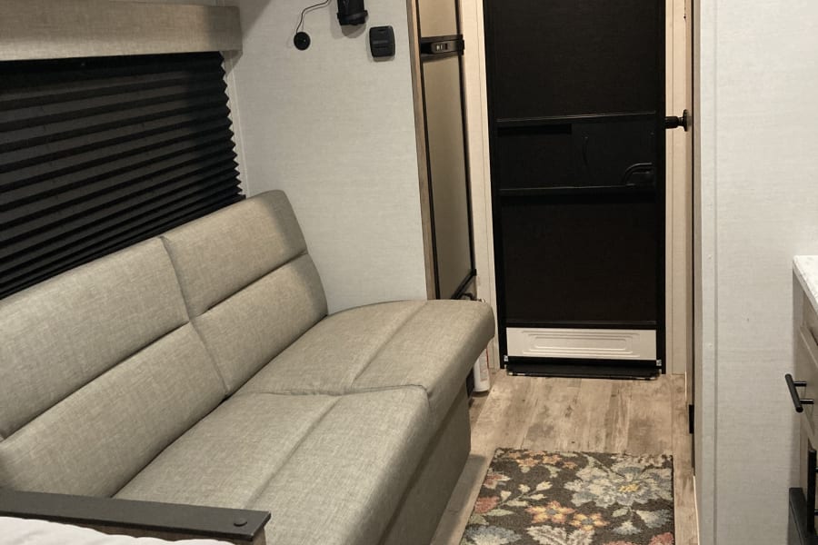 RV rental interior: light wood cabinets, grey sofa, window with blinds, TV, and door in a travel trailer living space.
