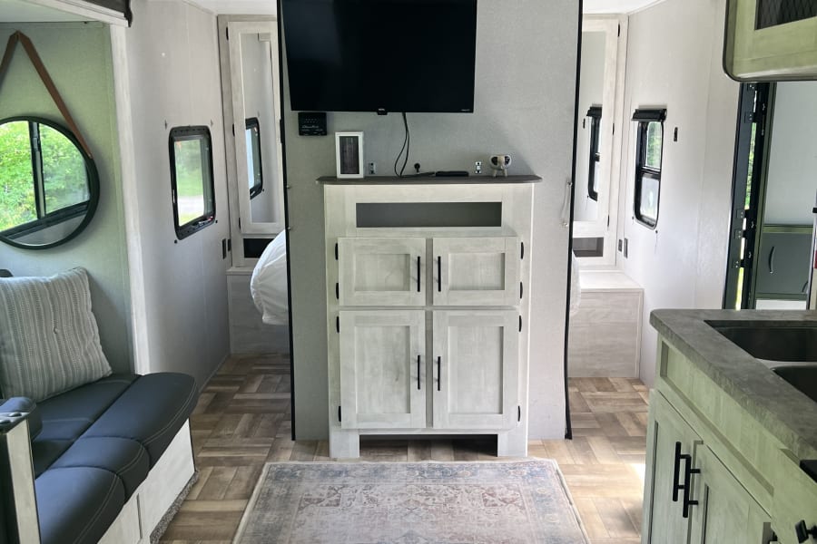 Modern RV rental interior: inviting sofa, central entertainment, kitchen, and rear bedroom hallway with wood floors.