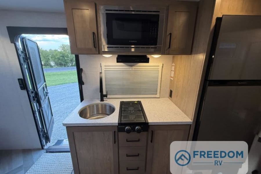 This RV rental kitchen interior features a sink, stove, microwave, fridge, and an open door showing the outdoors.