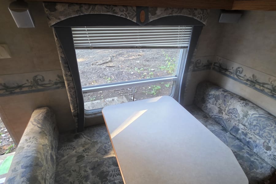 Cozy RV rental dinette: blue/gray patterned booth seating, wood cabinets, window showing outdoor view.