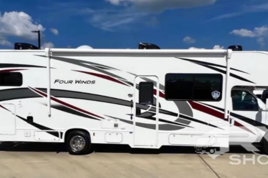 Side view of a white Four Winds Class C RV rental with black, red, and grey graphics, parked outdoors under a blue sky.