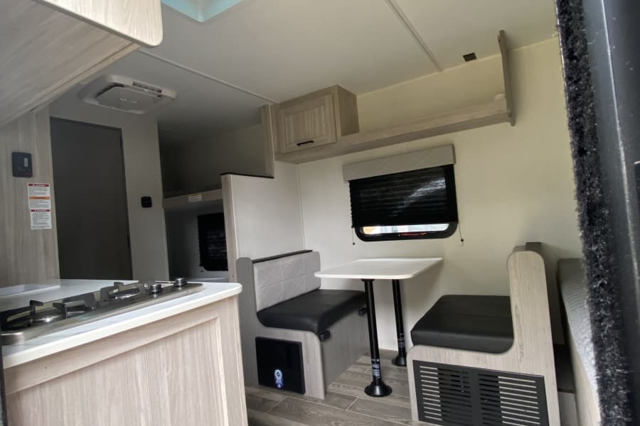 Modern RV rental interior: kitchen with stove, dining booth, wood-look floor, AC unit, and overhead storage.