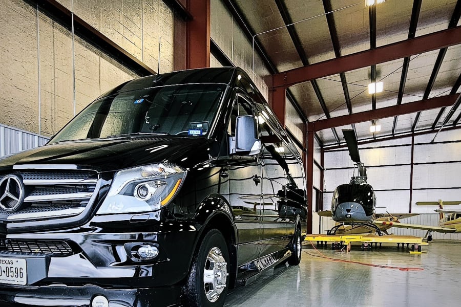 This sleek Mercedes Sprinter RV rental van stands ready in a hangar, sharing space with a helicopter & aircraft.