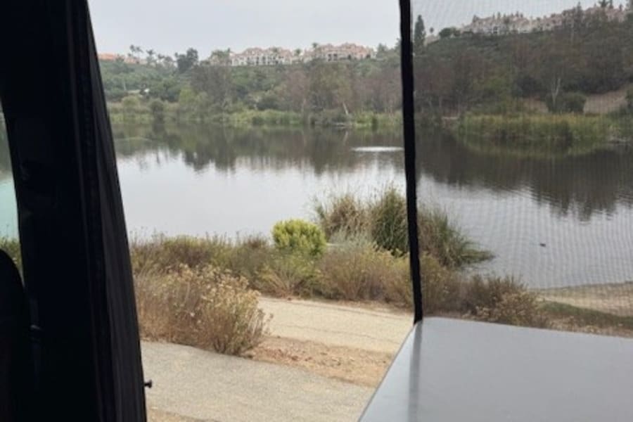 RV rental view: a tranquil lake reflecting an overcast sky, with distant homes and a path, seen through a screened window.