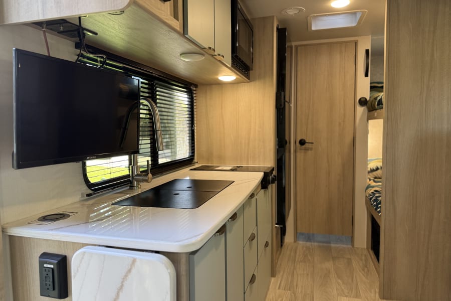 Modern RV rental interior features a kitchen with sink and stovetop, wall-mounted TV, and light wood design. Bright, functional space.
