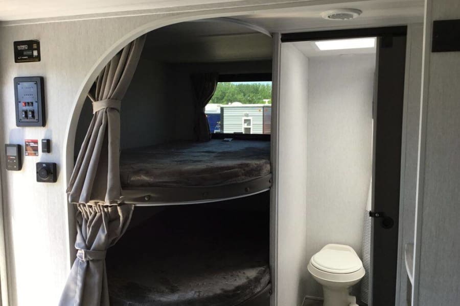 This RV rental interior features bunk beds with curtains, integrated controls, and a compact bathroom with a toilet.