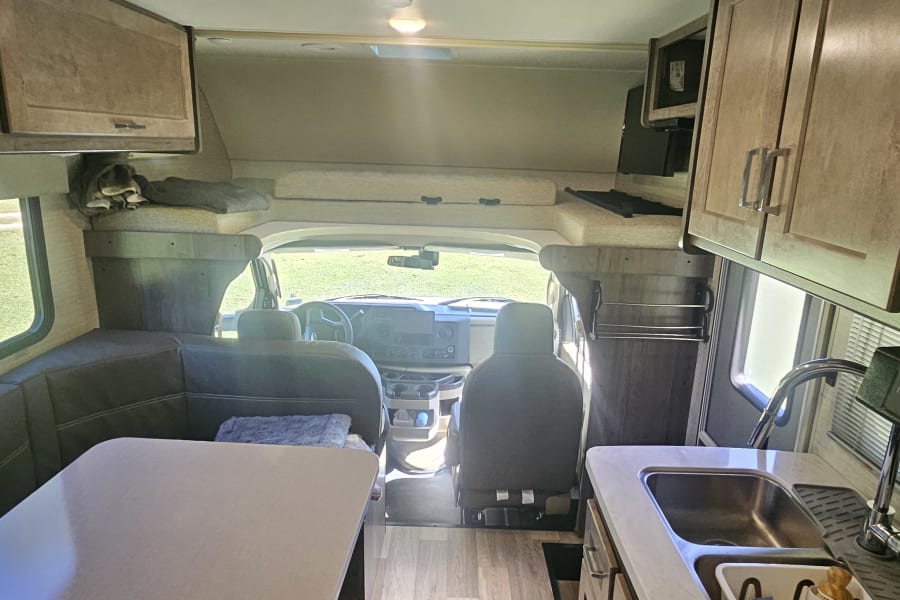 Inside this rv rental, a dinette, kitchenette with sink & microwave, driver's cab, and overhead bunk are visible.