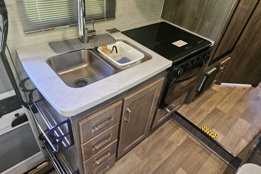 Inside an RV rental: a compact kitchen with a stainless steel sink, stove, wood-look flooring, and nearby seating.