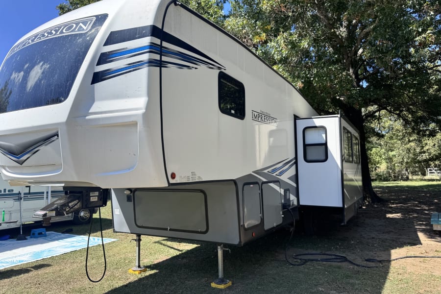 This white Forest River Impression fifth-wheel RV rental is set up at a shaded campsite, with its slide-out extended and leveling jacks deployed.