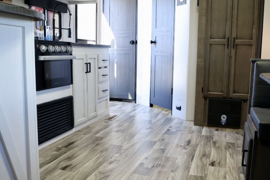 Bright RV rental interior with a modern kitchen, white cabinets, light wood-look flooring, and a glimpse of the dinette.