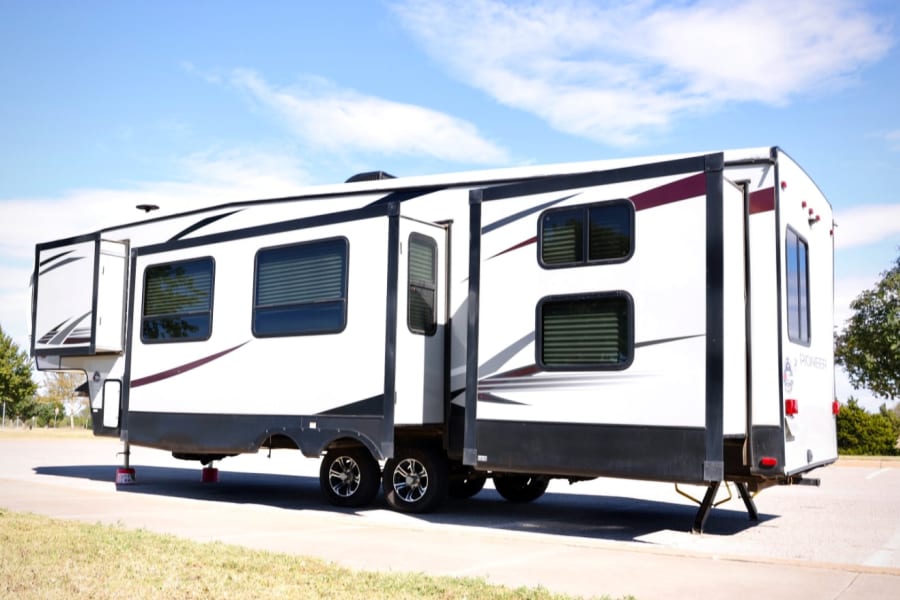 White fifth-wheel RV rental with extended slide-outs, parked and set up for camping under a clear blue sky.