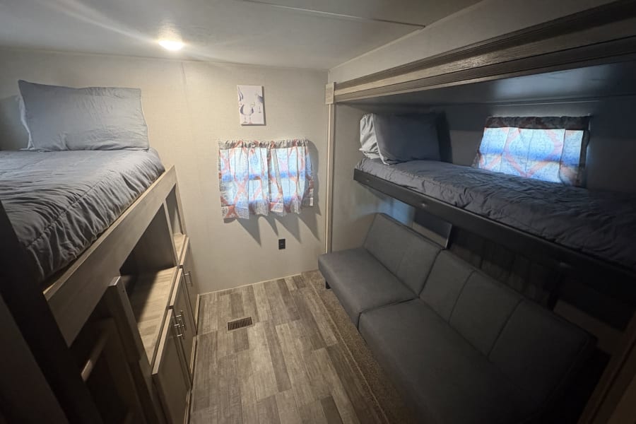 Inside an RV rental: a compact sleeping area with two sets of bunk beds, gray bedding, storage, and a sofa. Wood-look floor.