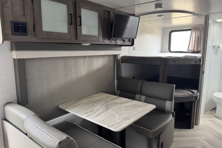 Modern rv rental interior: a dinette with marble-look table, gray seating, overhead cabinets, TV, and bunk beds.