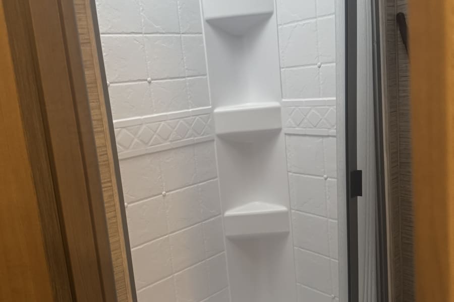 Inside an RV rental: a bright white shower with textured walls, corner shelves, and a translucent sliding door.
