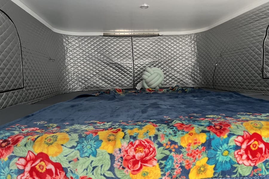 Cozy RV rental bedroom features a blue bed with a vibrant floral blanket, a light-green knot pillow, and quilted silver walls.