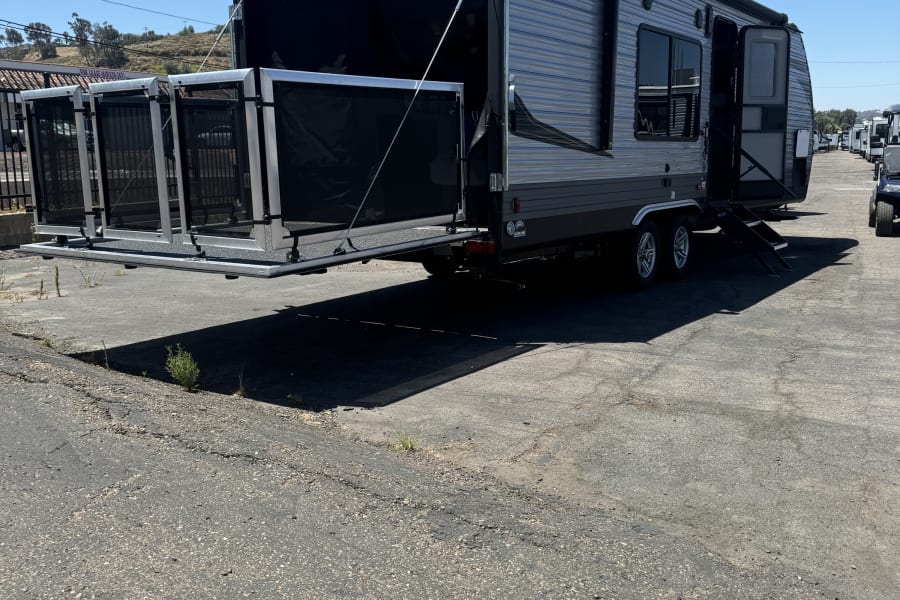 This grey RV rental, possibly a toy hauler, has its rear ramp lowered to form an outdoor patio deck, parked on asphalt under a clear sky.