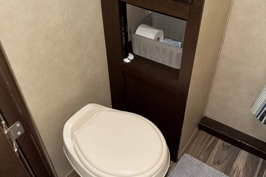 Compact RV rental bathroom with a toilet, cabinet storage for essentials, wood-look floor, and a gray rug.