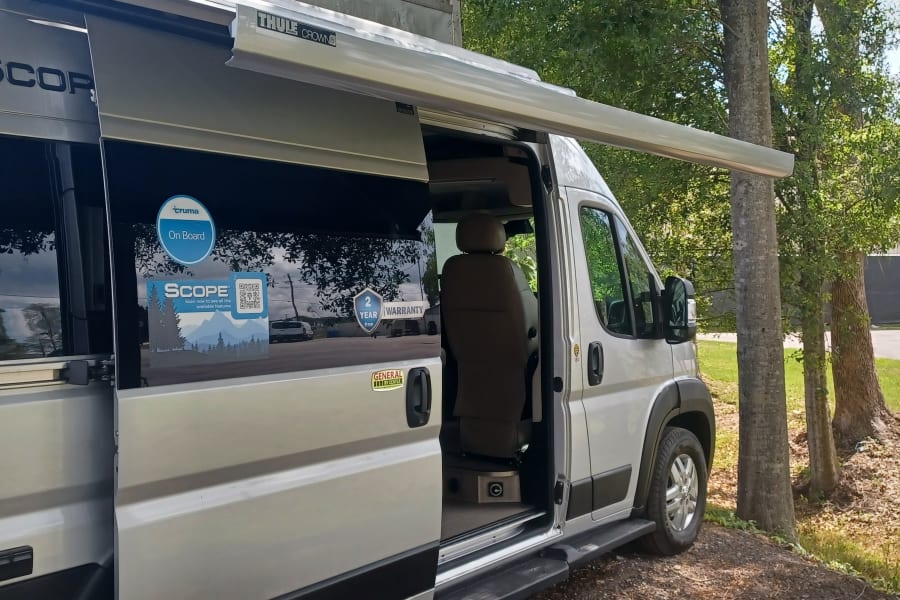 Silver **rv rental** van, branded 'SCOPE', with an extended awning and open side door, parked among trees.