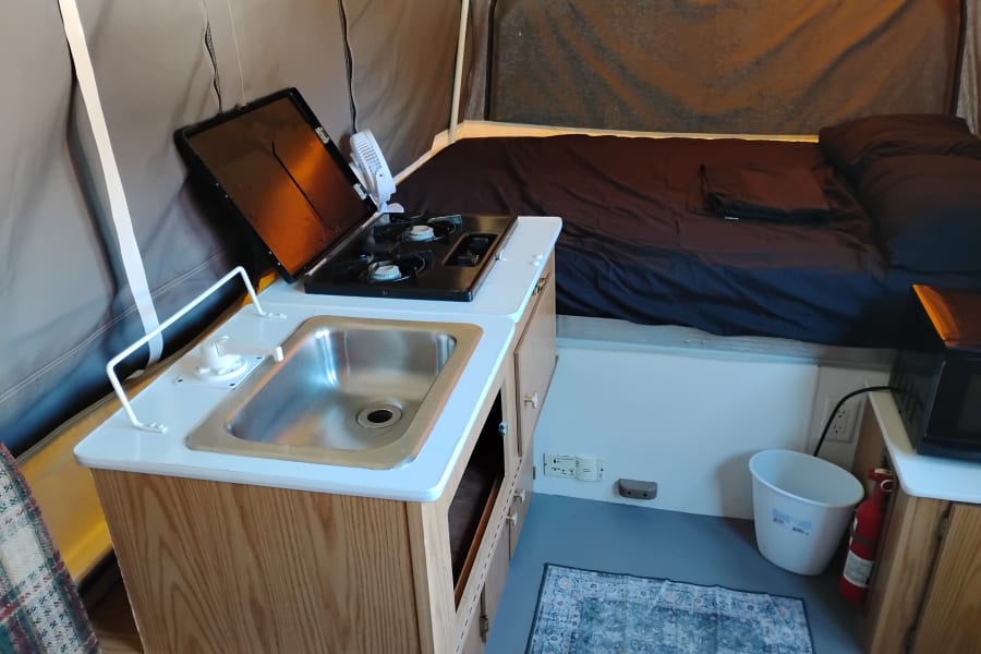 Inside an RV rental pop-up camper: a compact kitchenette with sink, stove, and fan sits opposite a made bed.