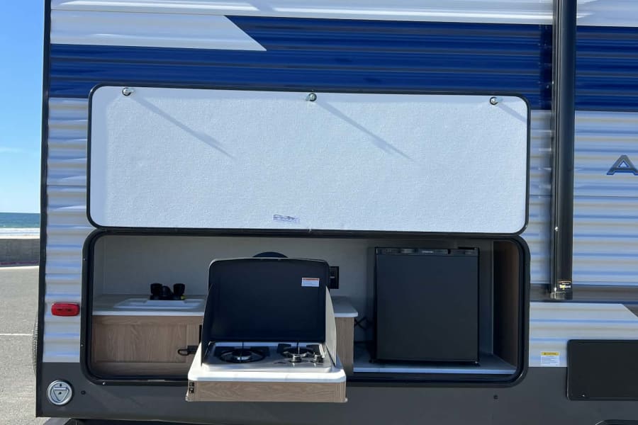 This RV rental features an open outdoor kitchen with a stove, sink, and mini-fridge, perfect for beachfront camping.
