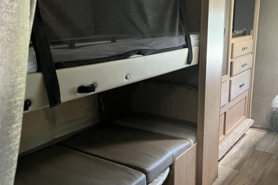 RV rental bunk beds: A cozy camper interior with a top bunk safety net, lower seating/bed, and adjacent storage/TV unit.