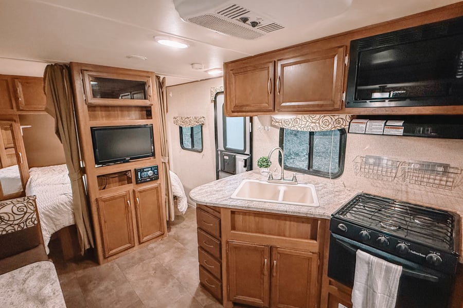 Cozy, well-maintained camper interior featuring a full kitchen with ample storage, a TV entertainment center, and a comfortable sleeping area separated by a curtain. Warm wood finishes and natural light create an inviting, homey feel.