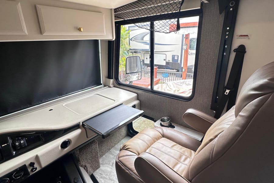 Spacious rv rental interior with a swiveled captain's chair, pull-out table, dashboard, and window view of other RVs.