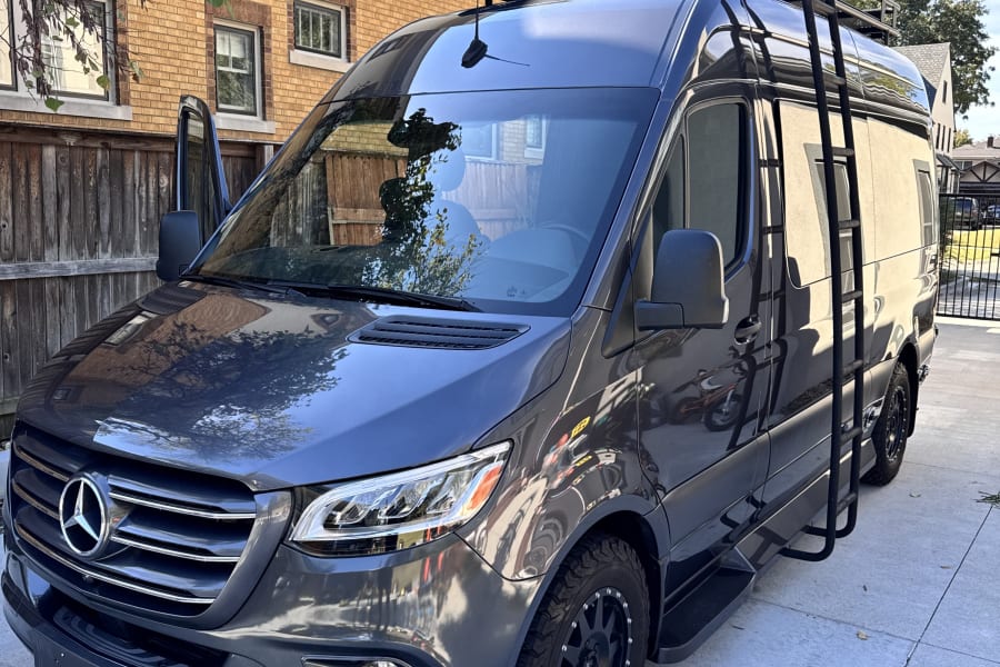 Custom dark gray Mercedes Sprinter RV rental with a roof rack, ladder, and off-road tires, parked on concrete.