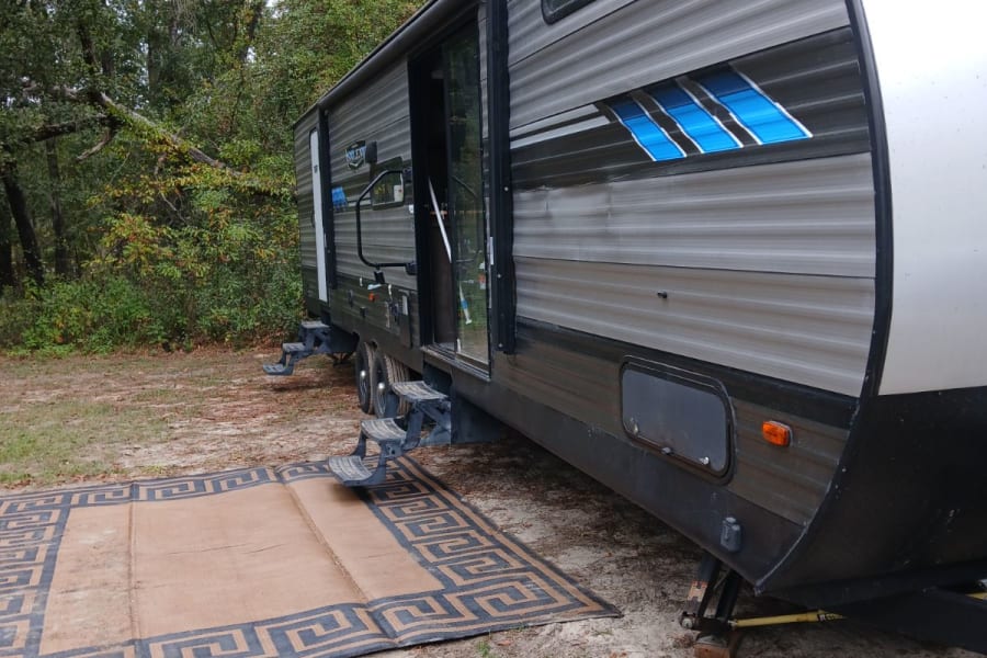 Travel trailer RV rental with steps & outdoor rug at a wooded campsite, ideal for nature exploration.