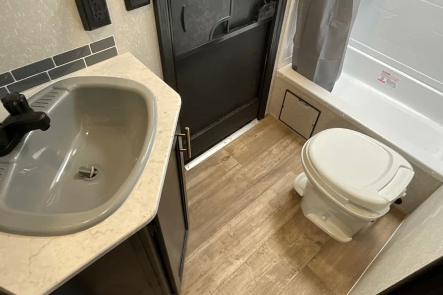 This RV rental bathroom shows a gray sink on a light counter, a white toilet, shower with curtain, and wood-look floor.