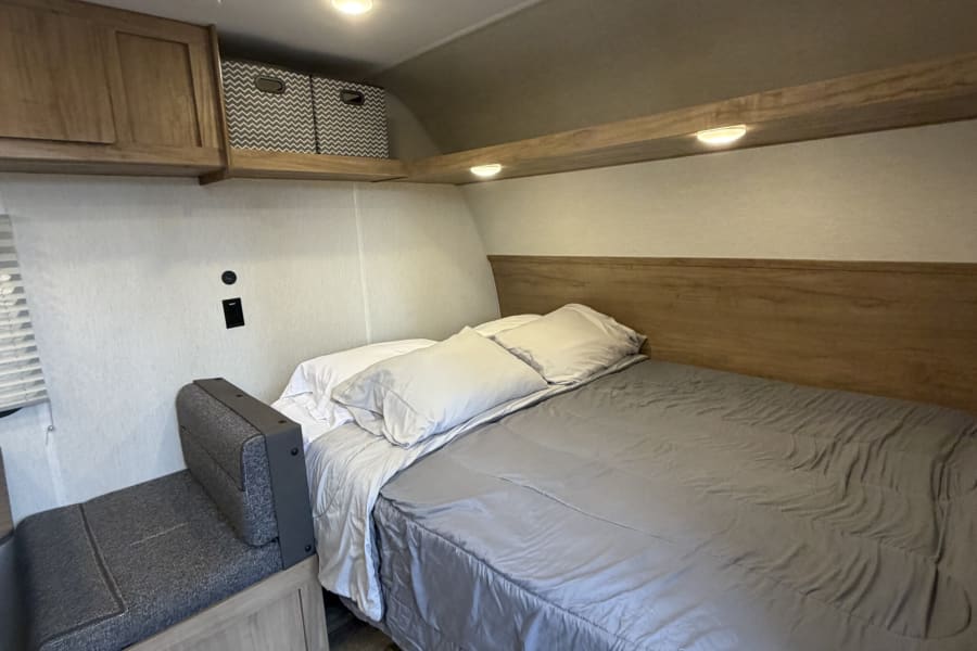 Cozy RV rental bedroom with a bed, gray bedding, small bench seat, overhead storage, and warm lighting.