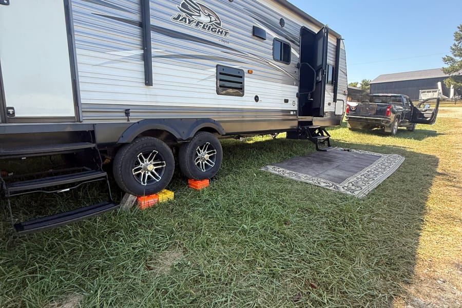 RV rental: Jay Flight travel trailer parked on grass with leveling blocks, steps, and an outdoor rug. Truck nearby.