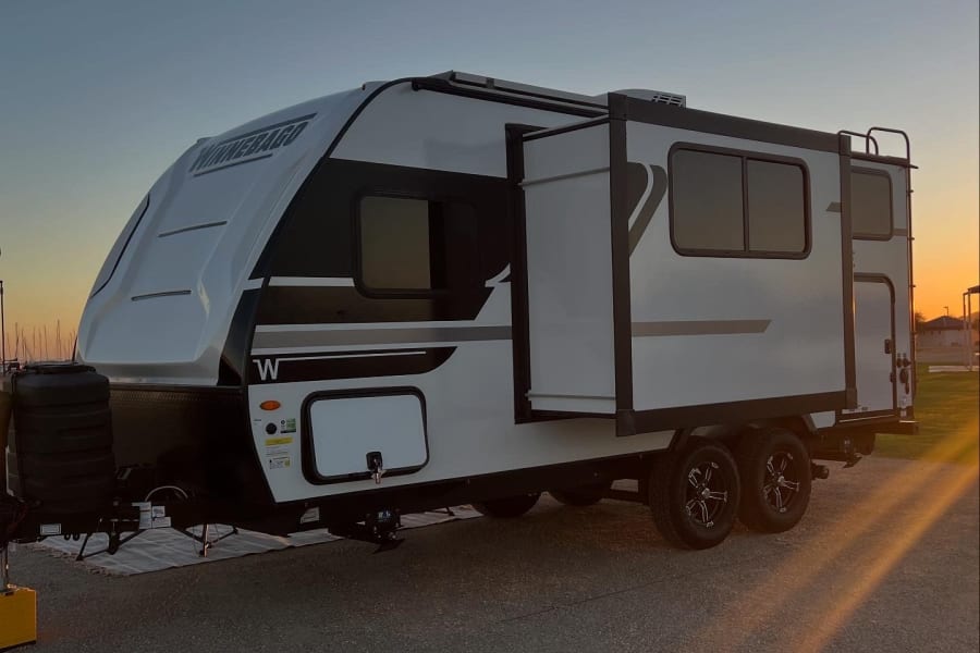 A white Winnebago travel trailer rv rental, with an extended slide-out, is parked at sunset. Perfect for outdoor adventure.