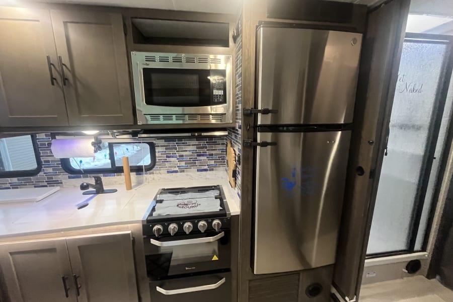 Modern RV rental kitchen: stainless steel fridge, microwave, gas range/oven, sink, blue-tiled backsplash, and wood cabinets.