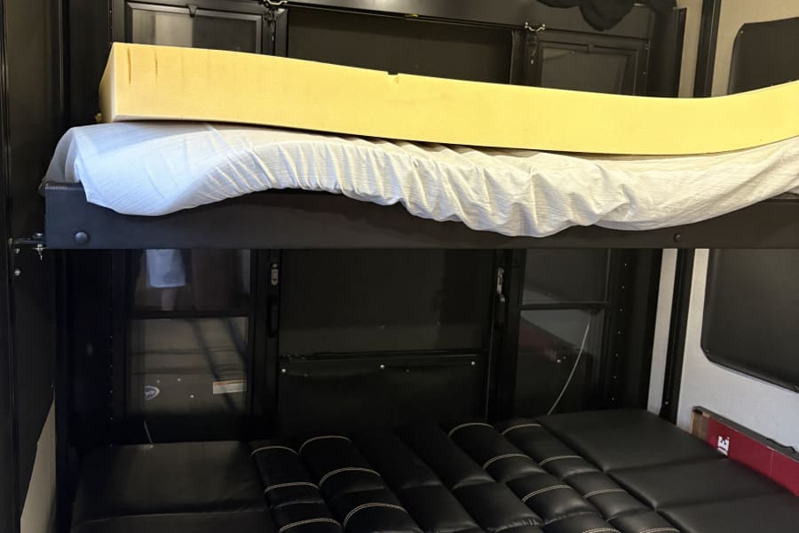 RV rental bunk beds in a toy hauler, featuring a top foam mattress and a lower black leatherette sofa-to-bed conversion.