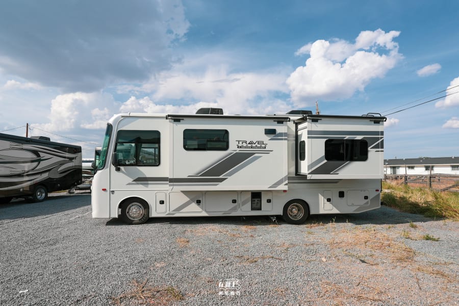 Modern white rv rental with its extended side section open, parked on a gravel lot under a partly cloudy sky.