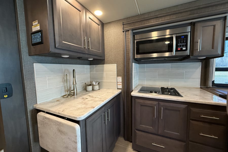 A modern and compact RV rental kitchen, featuring dark cabinets, light countertops, a sink, microwave, and stovetop.