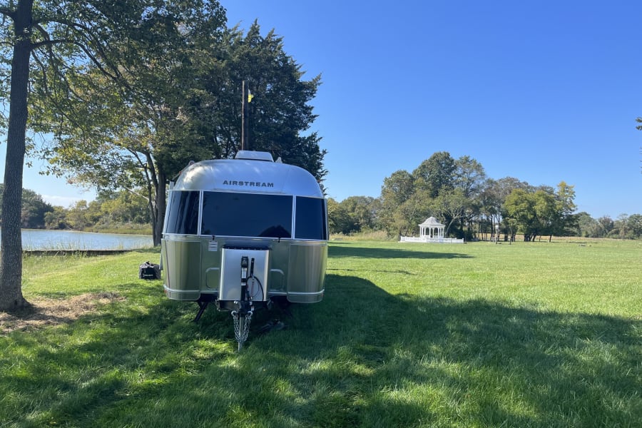 An Airstream RV rental is set up for a serene escape by a lake, with a white gazebo across the green field.
