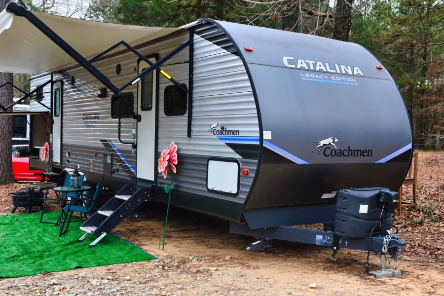 Coachmen Catalina rv rental at a wooded campsite, featuring an awning, outdoor seating, and fire pit.
