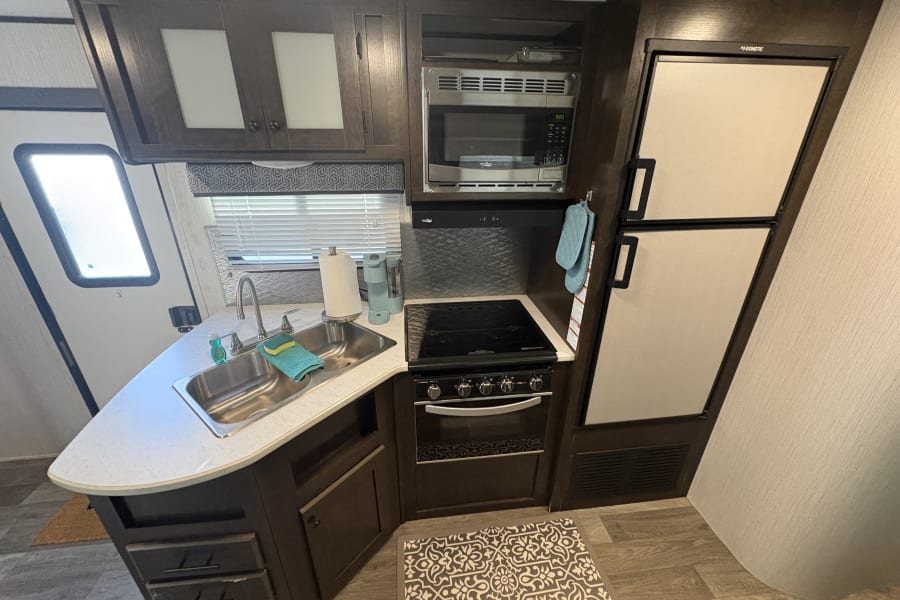 Modern RV rental kitchen. Features include a double sink, microwave, Dometic fridge, stove, and dark wood cabinets for travel.