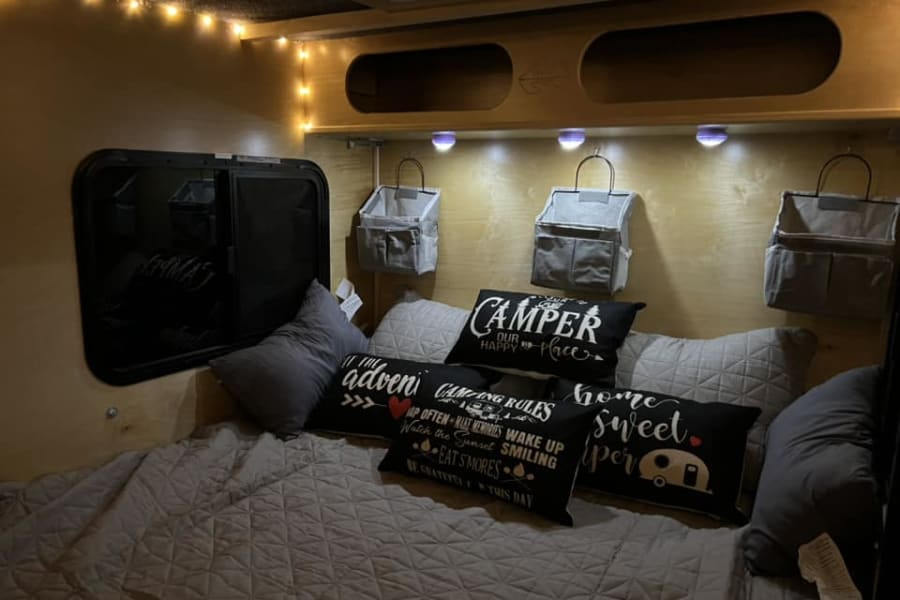 Inviting RV rental interior: a comfy bed with "Camper Our Happy Place" pillows, ambient lights, and a window for camping adventures.