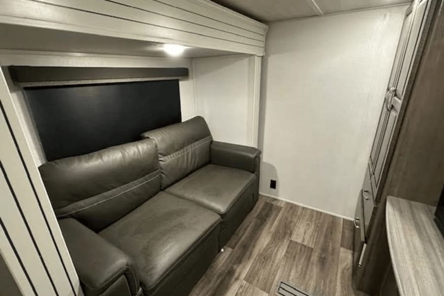 Dark gray sofa, window blind, and storage cabinet in a clean RV rental interior with wood-look flooring.
