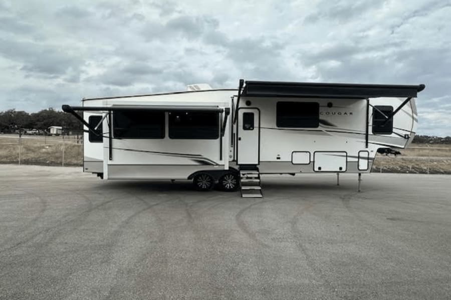 This spacious white Cougar fifth-wheel RV rental, with extended slides and deployed awning, awaits your next getaway under cloudy skies.