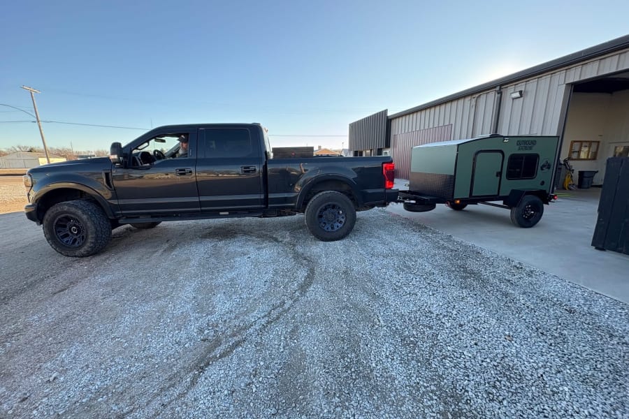 A dark truck tows an Outbound Extreme camper on gravel, ready for your RV rental adventure.