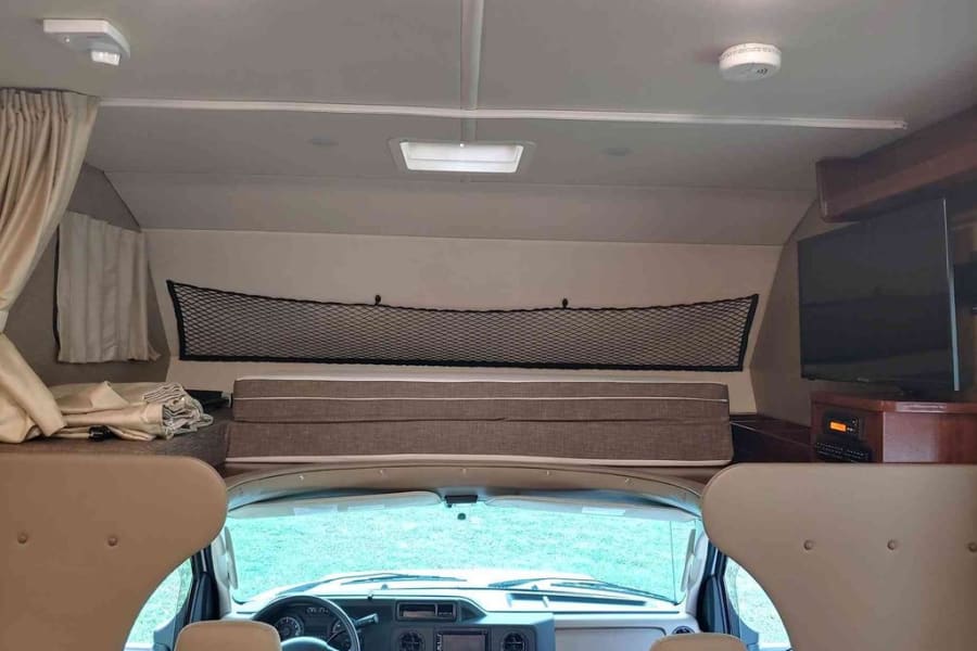 **RV rental** interior: Front view showing cab-over bunk with safety net, cockpit, TV, and ceiling safety detector.