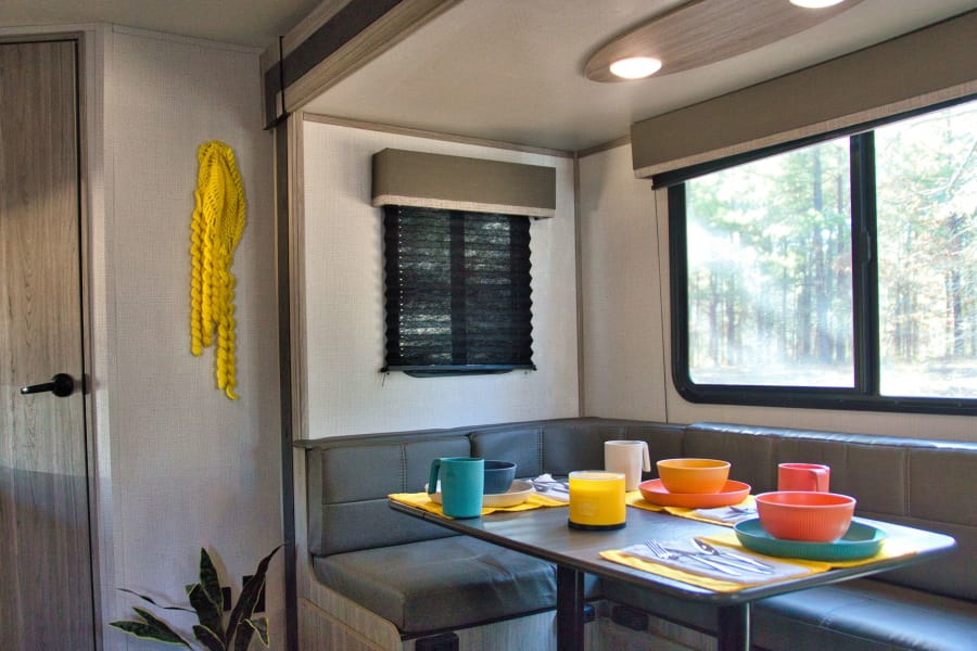 Cozy RV rental dinette interior: booth seating, colorful dishes set on table, bright windows with tree views, and a plant.