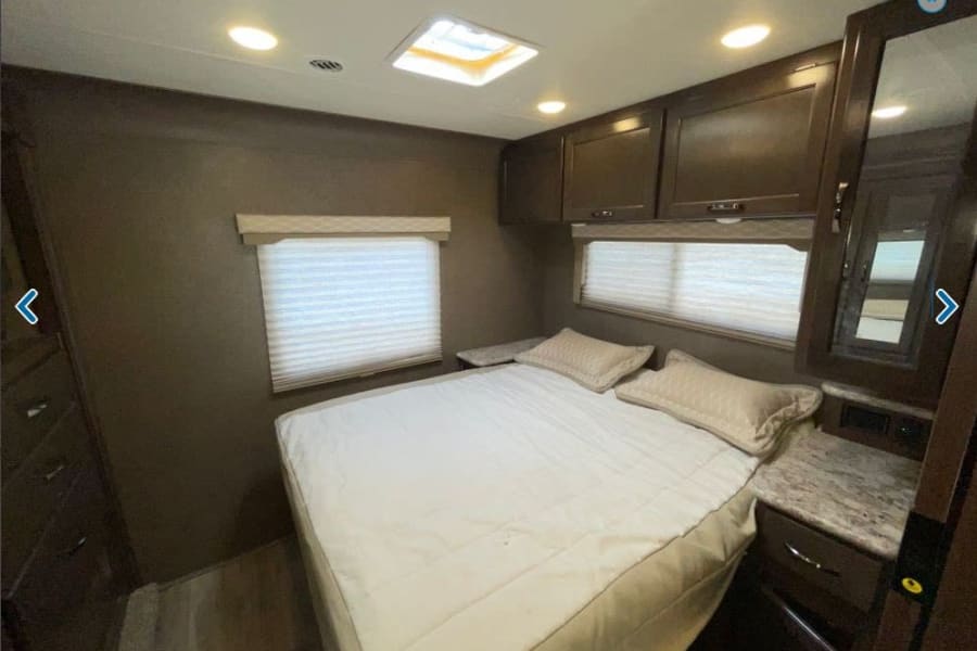 Inviting RV rental bedroom with a comfortable bed, two windows with blinds, and practical overhead storage cabinets.