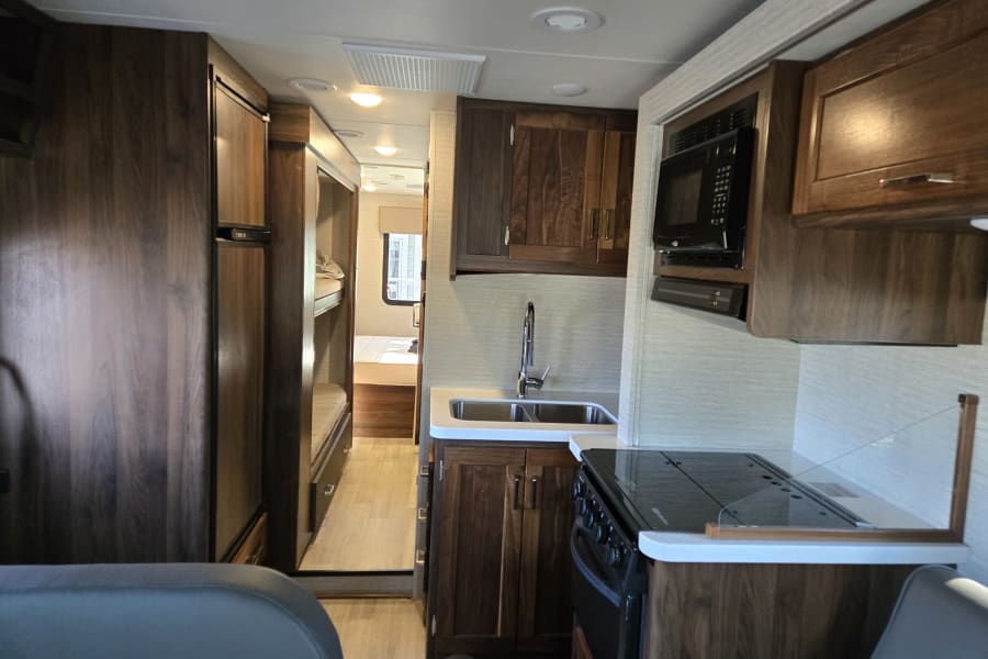 RV rental interior: kitchen with sink, stove, microwave, fridge, and a hallway leading to bunk beds and a bedroom.